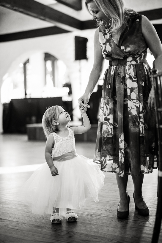 A toddler in a white dress looks up to her mother in an empty reception room. The picture was taken by a Denver wedding photojournalist at The Villa Parker