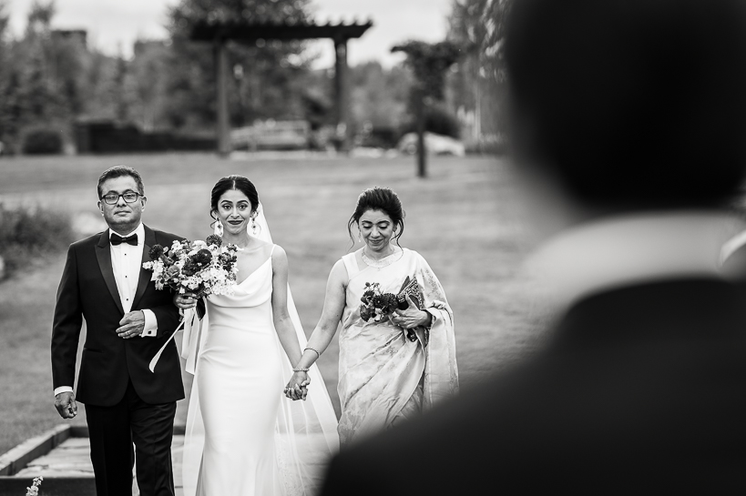 A bride walks into an outdoor wedding ceremony accompanied by her parents, as seen from the perspective of the groom.