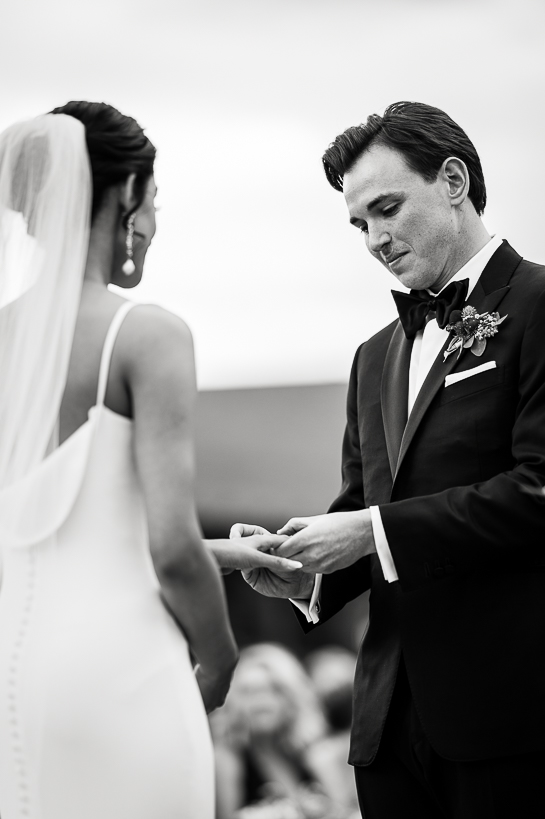 Denver black and white wedding photograph of a groom slipping a ring on a bride's finger during an outdoor ceremony.