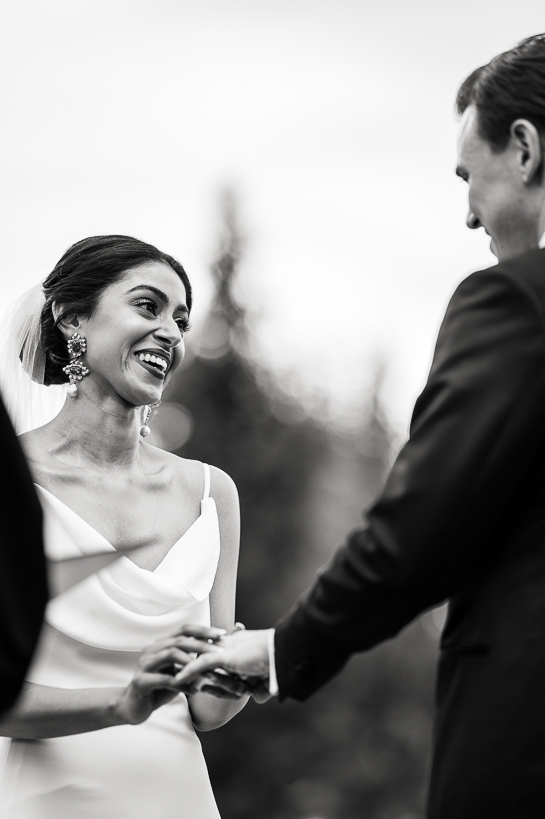 Black and white vertical image of bride slipping a ring on her groom's finger during an outdoor wedding ceremony in the Colorado mountains.