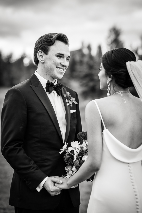 A groom and bride talk in an open field with pine trees in the background in a black and white photograph.