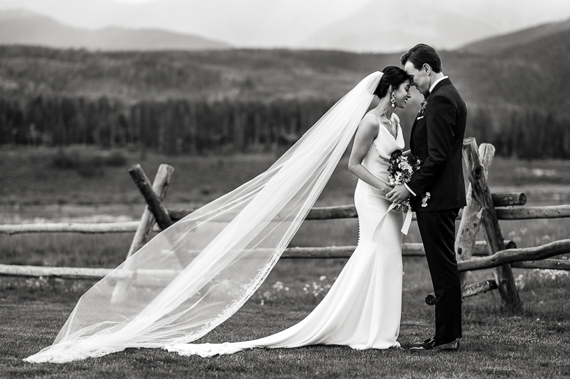 A bride and groom touch foreheads in a tender moment, her long veil blowing behind her in the breeze. It is a rural setting, near an old wooden fence, with a large field, then pine trees and mountains in the distance.