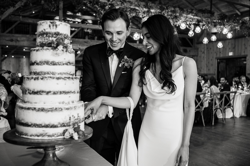 In image by Denver wedding photographer, a bride and groom cut a multi-tiered cake in a black and white image.